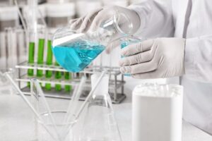 clinical-worker-dressed-white-gown-gloves-holding-glass-beakers-with-blue-liquid_273609-13421